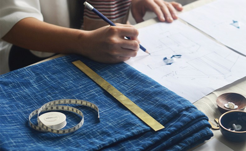 A person's hand holds a pencil over a blue patterned fabric spread on a table. A blueprint with faint blue markings lies beneath the fabric. To the left, a tape measure and a gold ruler are visible, along with a roll of white tape. Two small bowls, one containing a white object and the other with a blue object, are also on the table.