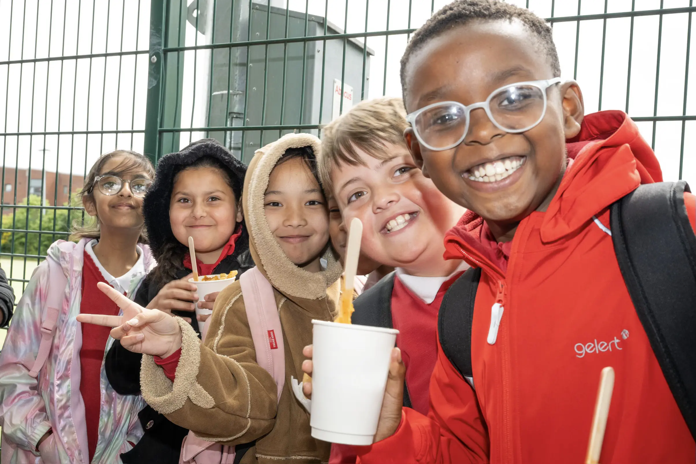 A group of smiling schoolchildren standing outside next to a fence, holding warm meals and posing cheerfully.