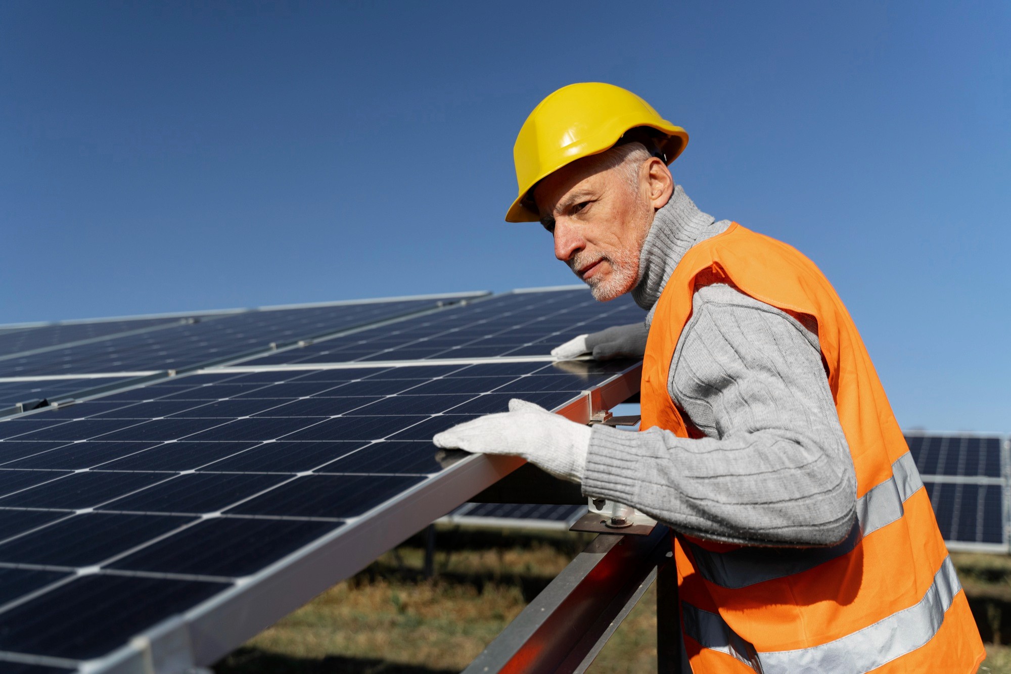 A worker in a hard hat and safety vest inspects solar panels under a clear blue sky.