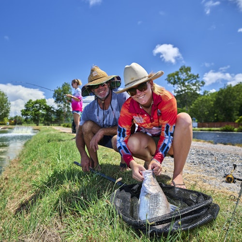 两个人戴着帽子在池塘边跪下，微笑着用网子捧着一条鱼。在背景中，另一个人在钓鱼。