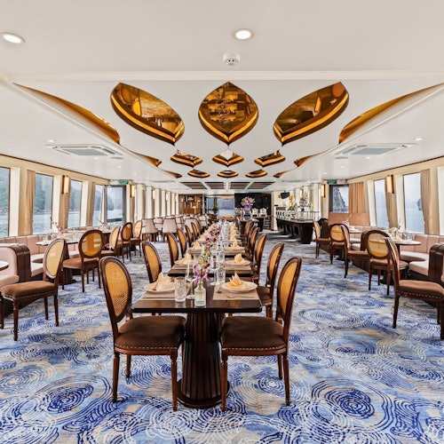 En elegant spisestue med et blå hvirvlende mønstret tæppe, træborde og brune polstrede stole, med gyldne loftsdetaljer.