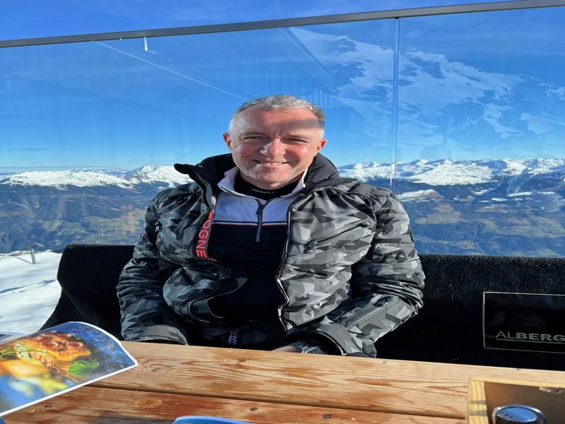 A person wearing a camo jacket sits at an outdoor wooden table, surrounded by snow-covered mountains and clear blue skies, with a view enhanced by a glass barrier.