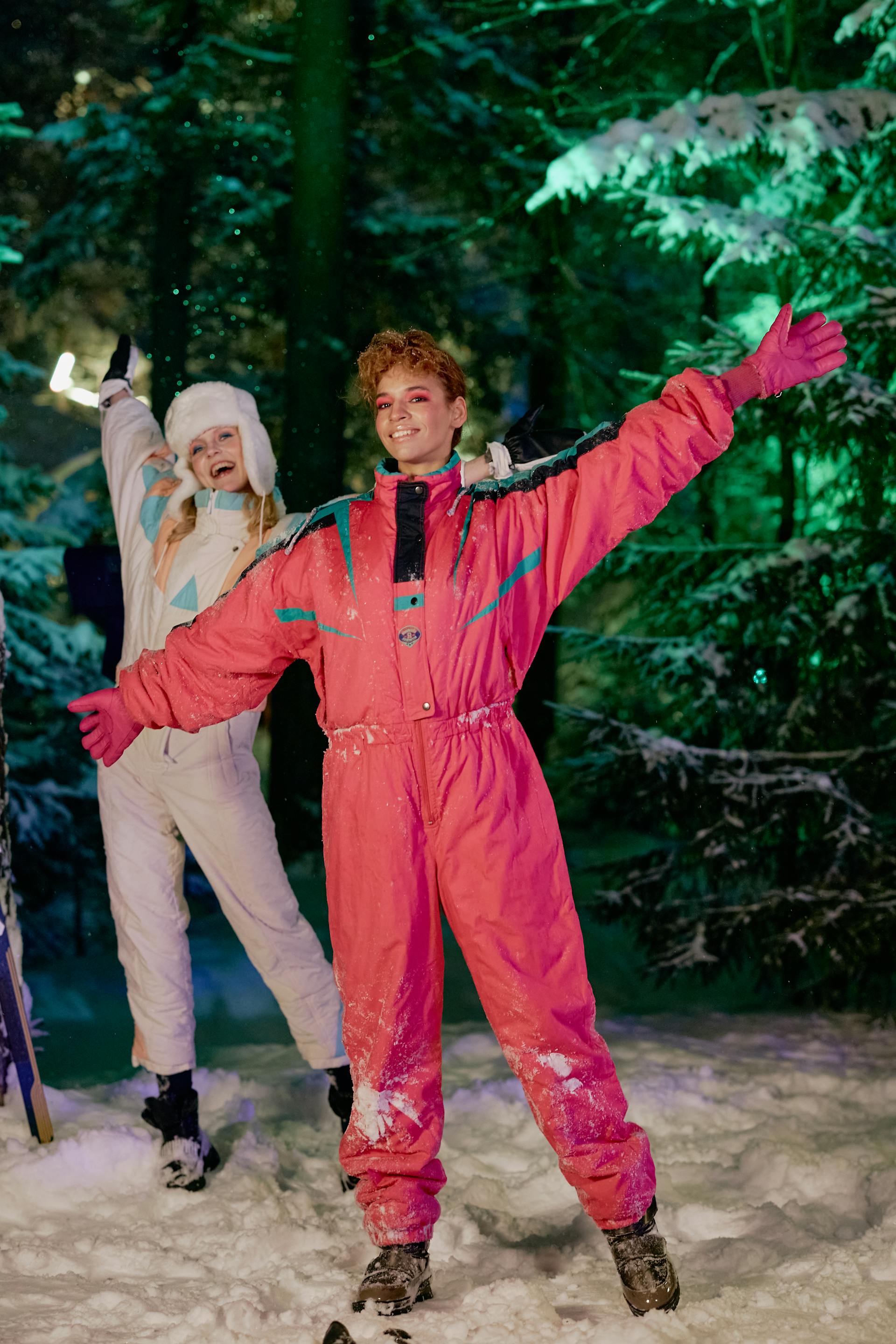 A person in a bright pink one-piece ski suit stands with arms outstretched in a snowy forest at night, while another person in a white suit waves in the background.