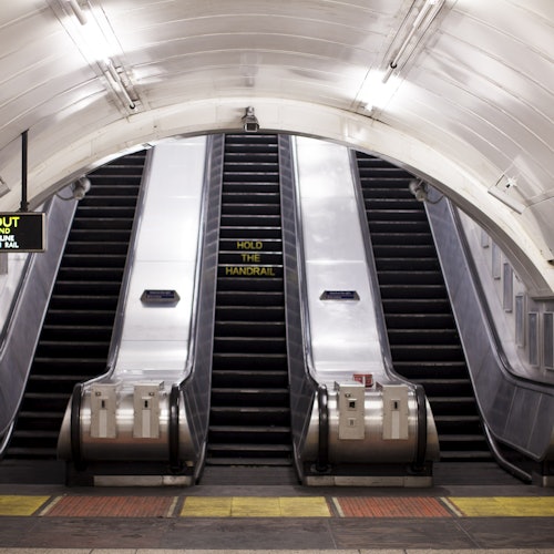 一个空地铁站的自动扶梯，中间有一个“扶好扶手”的标志。上方是白色瓷砖拱门。