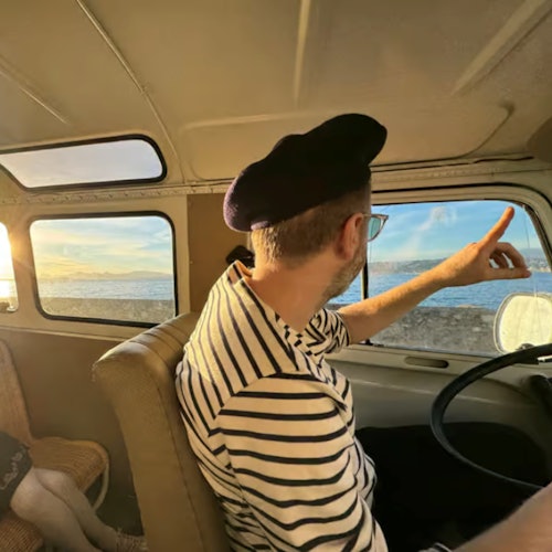 En person i stripete skjorte og beret peker ut av et varebilvindu mot havet, opplyst av den nedadgående solen.