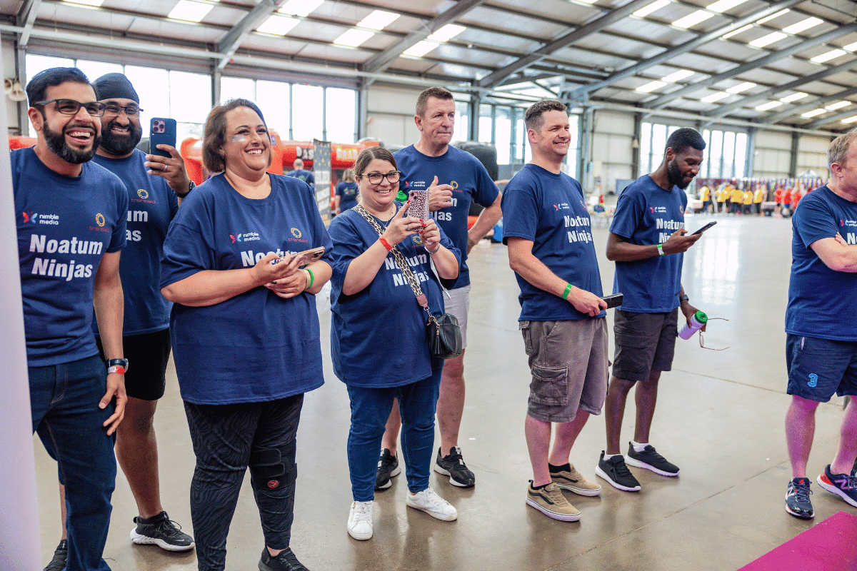 Outdoor team-building event with participants wearing matching blue shirts, taking part in inflatable obstacle courses and games, smiling and cheering, with the words 'ENTER A TEAM' in large bold white and green text across the image.