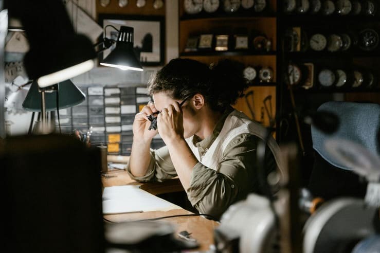 A woman sitting at a desk in a bright office, working on a laptop with notebooks, a cup, and a desk lamp beside her.