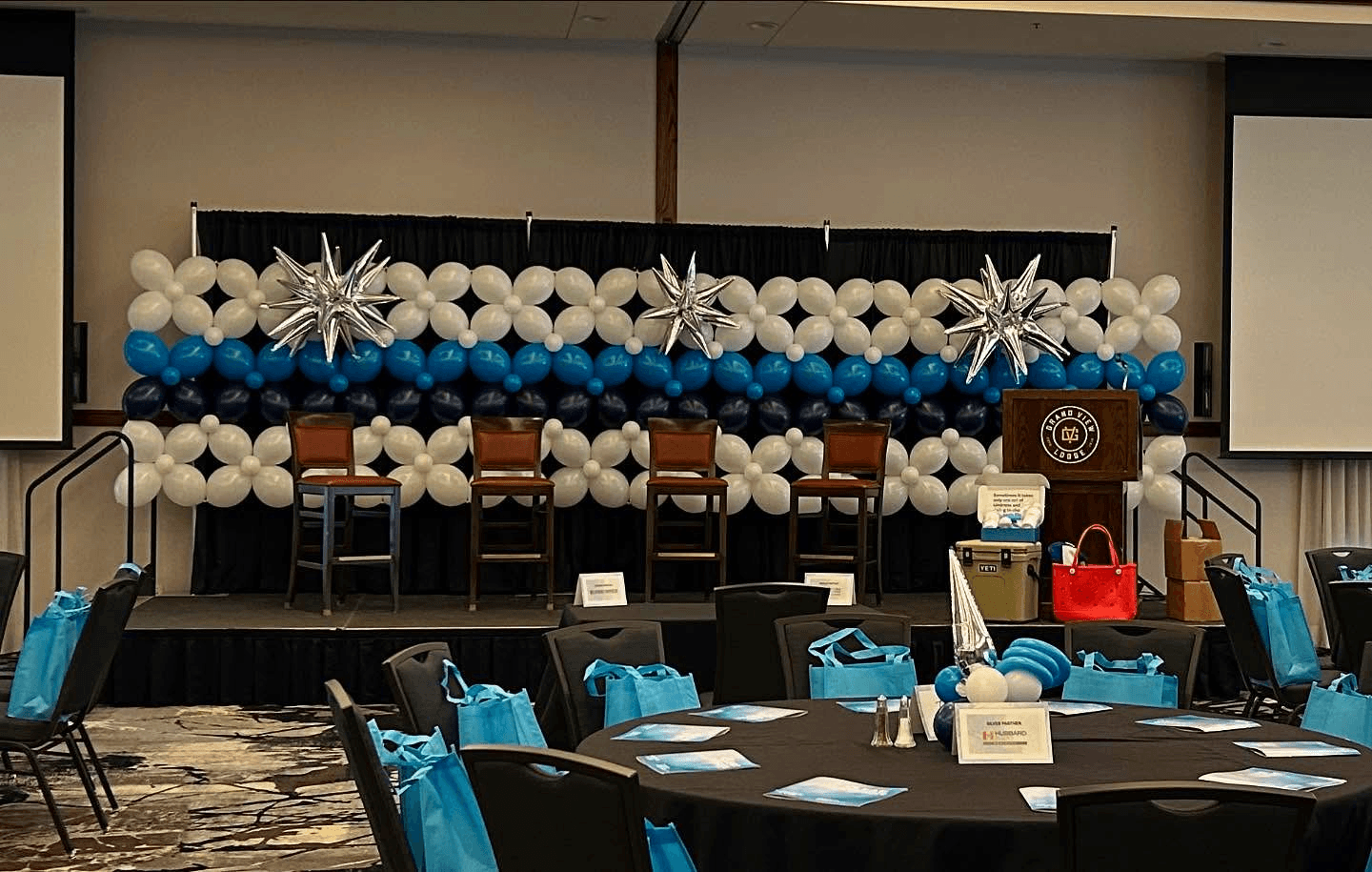 The image shows a conference room set up with neatly arranged chairs on a stage, framed by a vibrant wall of white and blue balloons, including decorative star-shaped balloons, with tables in the foreground covered in black tablecloths and adorned with blue gift bags.