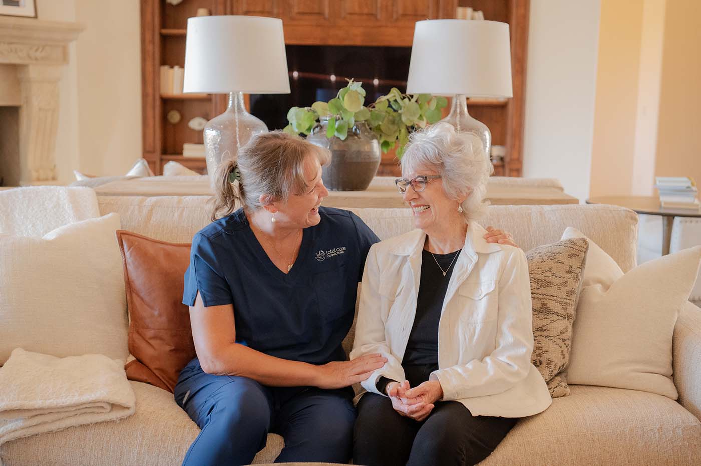 Two women sitting on a couch, one wearing blue scrubs and the other wearing a white jacket, smiling at each other.