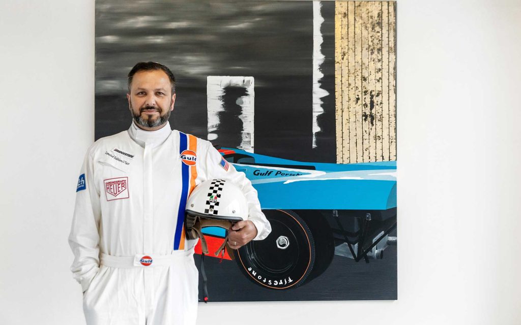 A man in car racing gear standing in front of a painting.