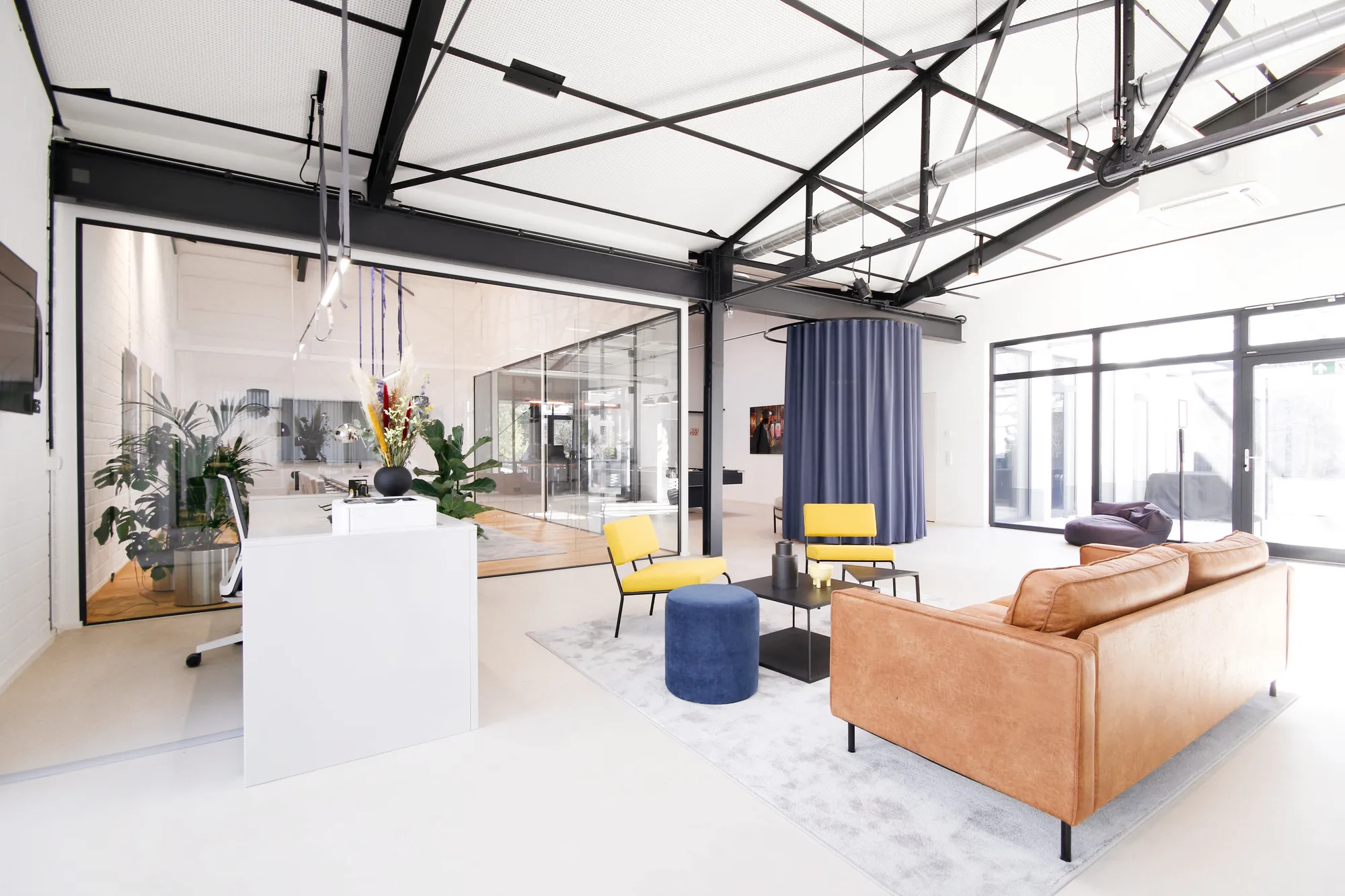 Open loft with leather sofa, yellow chairs, blue pouf and a glass-walled meeting room.