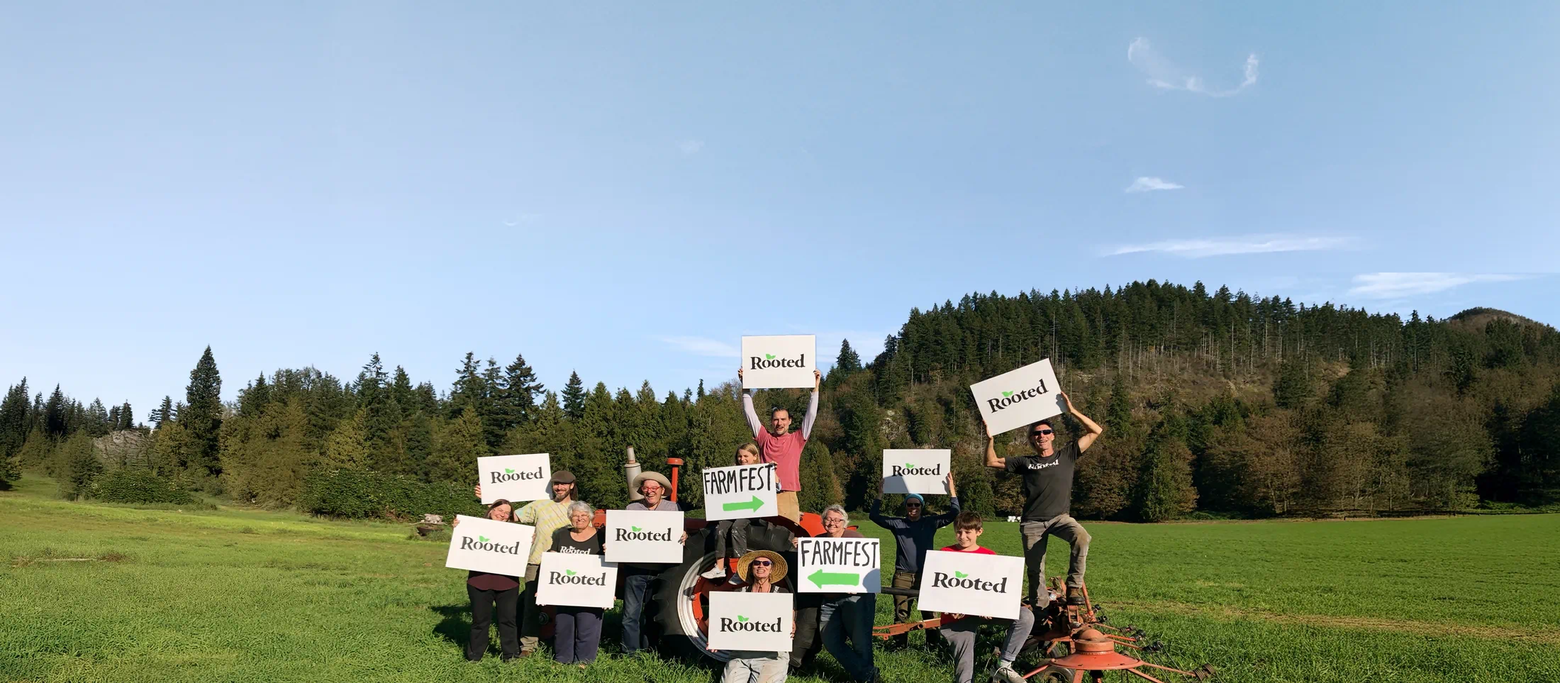 Group of neighbors standing in a grassy field holding “Rooted” and “FarmFest” signs during a community event.