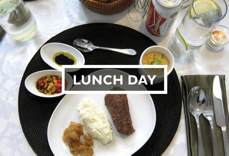 A black placemat holds a white plate with three food items: mashed potatoes, a piece of meat, and sliced carrots. Beside the plate are three small white bowls containing different sauces or condiments: dark sauce, a yellow sauce with green herbs, and a small red sauce. A fork and knife rest on a dark green napkin to the right of the plate. Two glasses of water and a can of Budweiser beer are visible on the table. A spoon and bowl are placed to the left of the plate, and a small woven basket is partially visible at the top of the image. The tablecloth has a subtle light pattern.