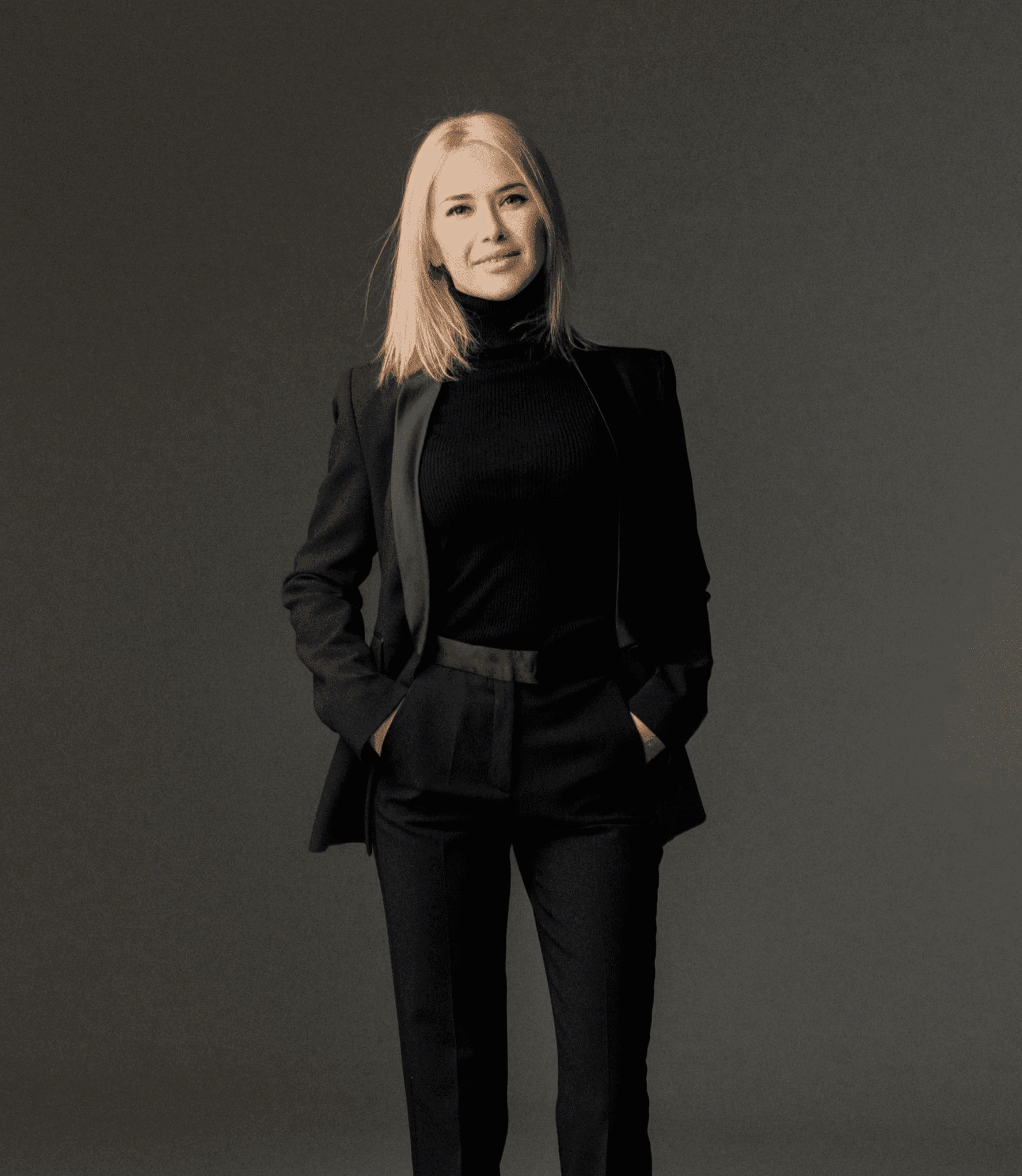 A confident person in a stylish black suit with hands in pockets stands against a neutral backdrop, exuding professionalism and elegance.