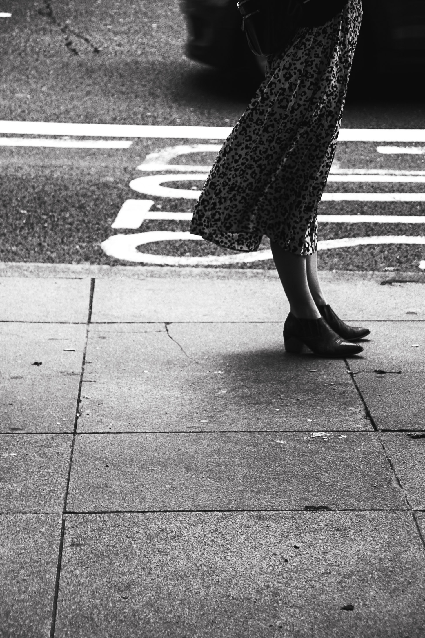 Black and white street photograph of a woman standing on a sidewalk, her shadow cast on the pavement.