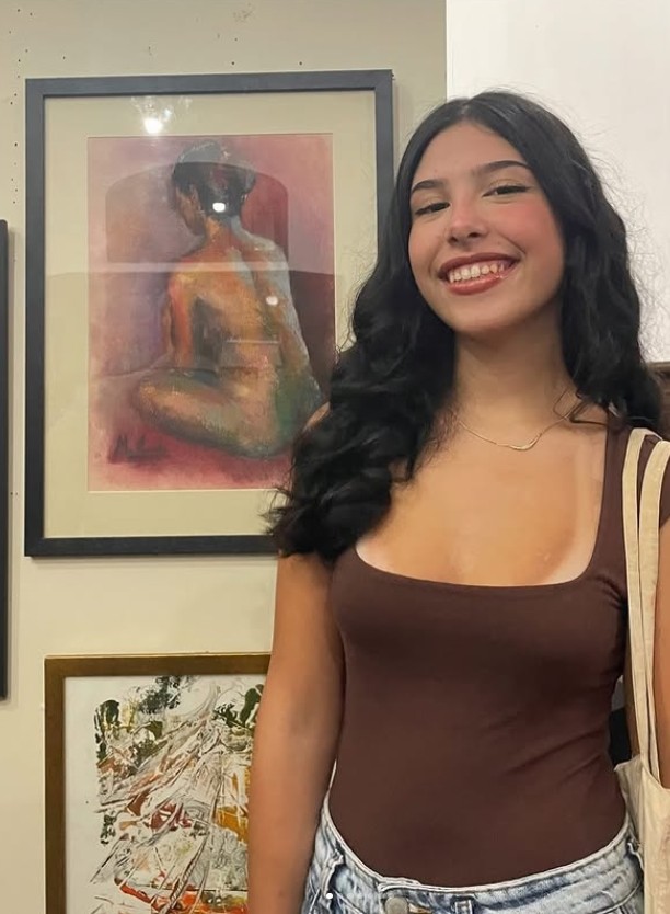 Menina jovem de cabelos pretos ondulados, sorrindo em frente a um quadro 