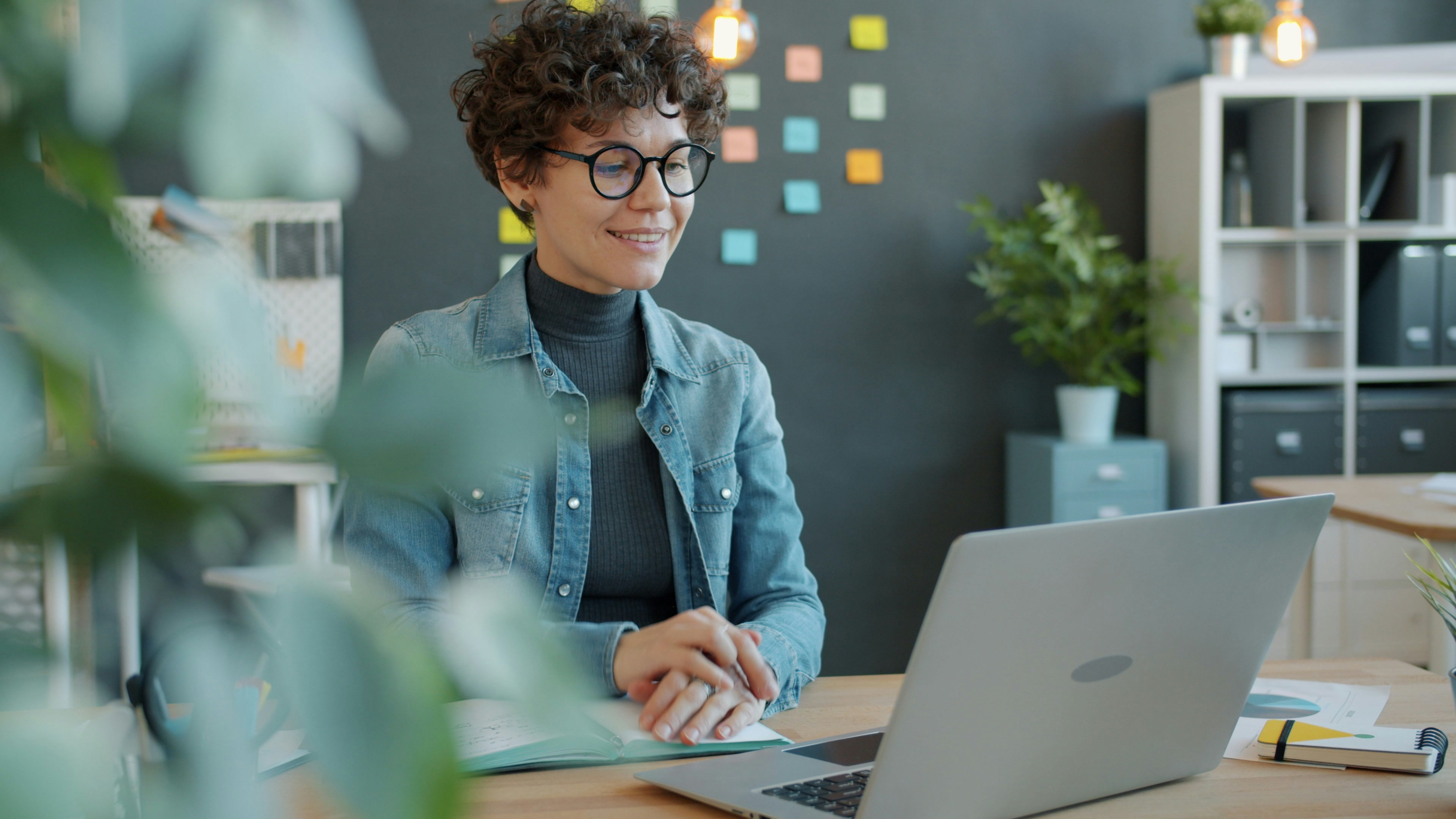 A smiling woman with curly hair works at a laptop.
