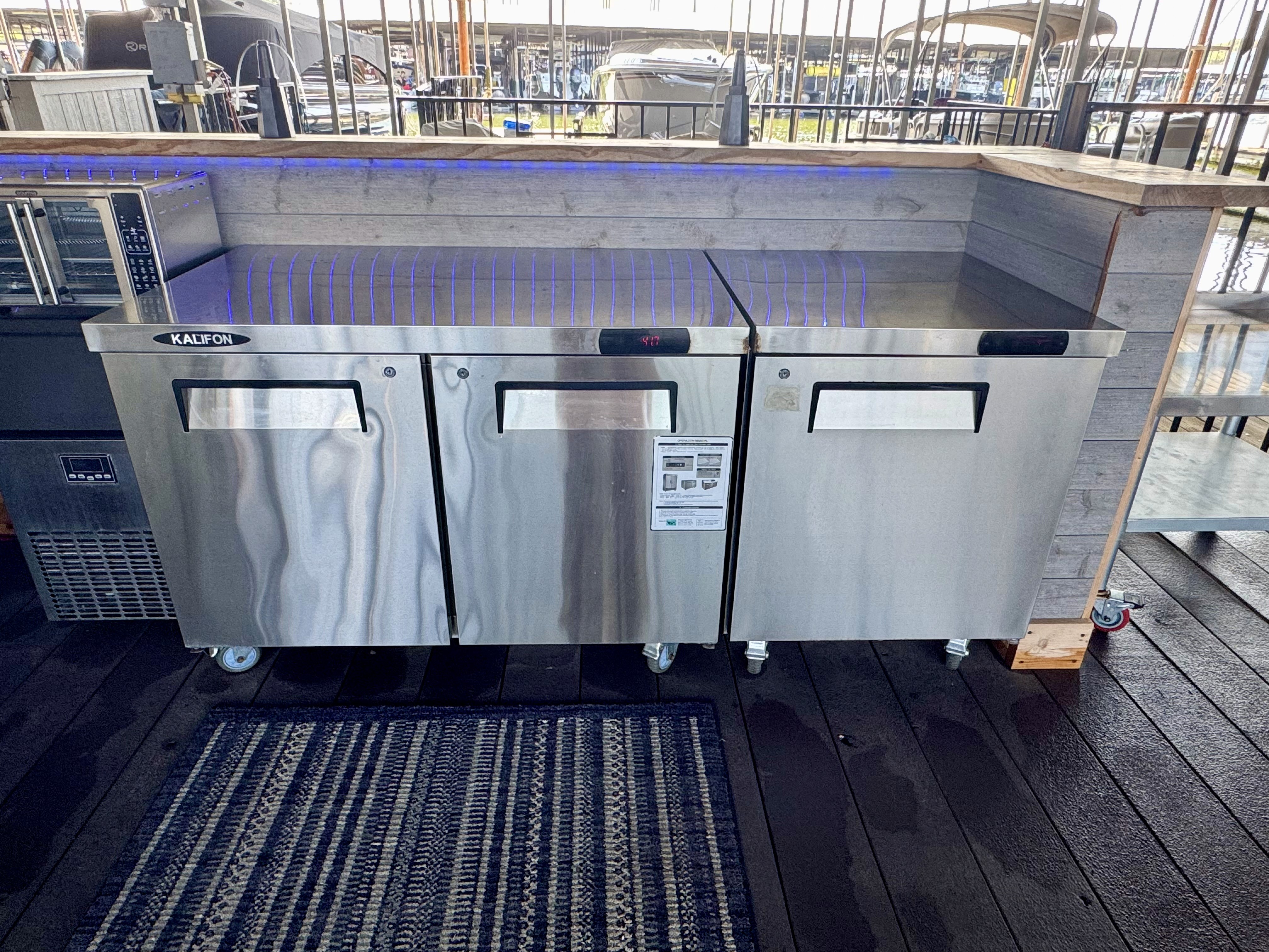 The image shows a sleek, stainless steel outdoor commercial kitchen setup, featuring two large refrigeration units under a rustic wooden countertop, ideal for professional use in dining establishments or catered events.