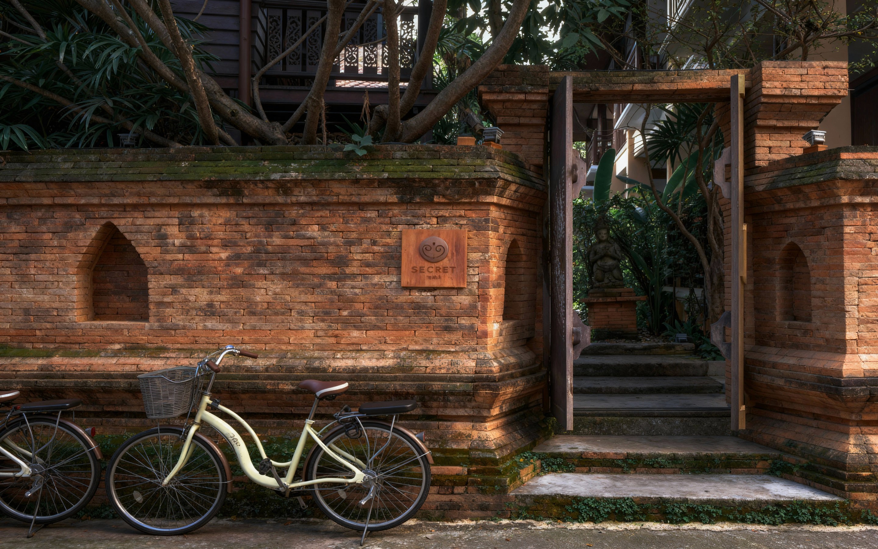 Rustic red-brick wall and open wooden gate leading into a lush garden courtyard in Chiang Mai’s Old City, with two vintage bicycles parked in front and a stone statue visible through the doorway.