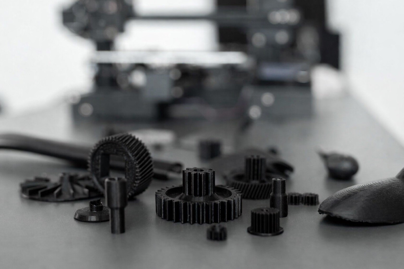 A collection of various black 3D-printed mechanical components, including spur gears, ring gears, and small cylindrical spacers, scattered across a dark gray surface. The parts feature a matte finish with visible fine layer lines. In the background, a desktop 3D printer is visible but heavily blurred, creating a shallow depth of field that keeps the focus on the finished hardware.