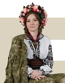 Ветеранка в українському національному вбранні та квітковому вінку сидить із накинутим на плечі військовим кітелем.
