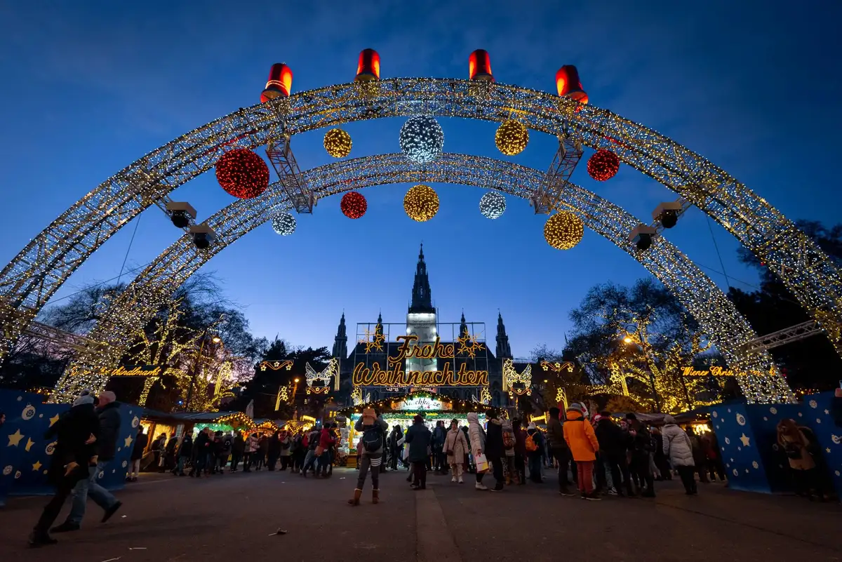 A large illuminated arch decorated with oversized ornaments frames a lit Christmas market in front of a castle at dusk.