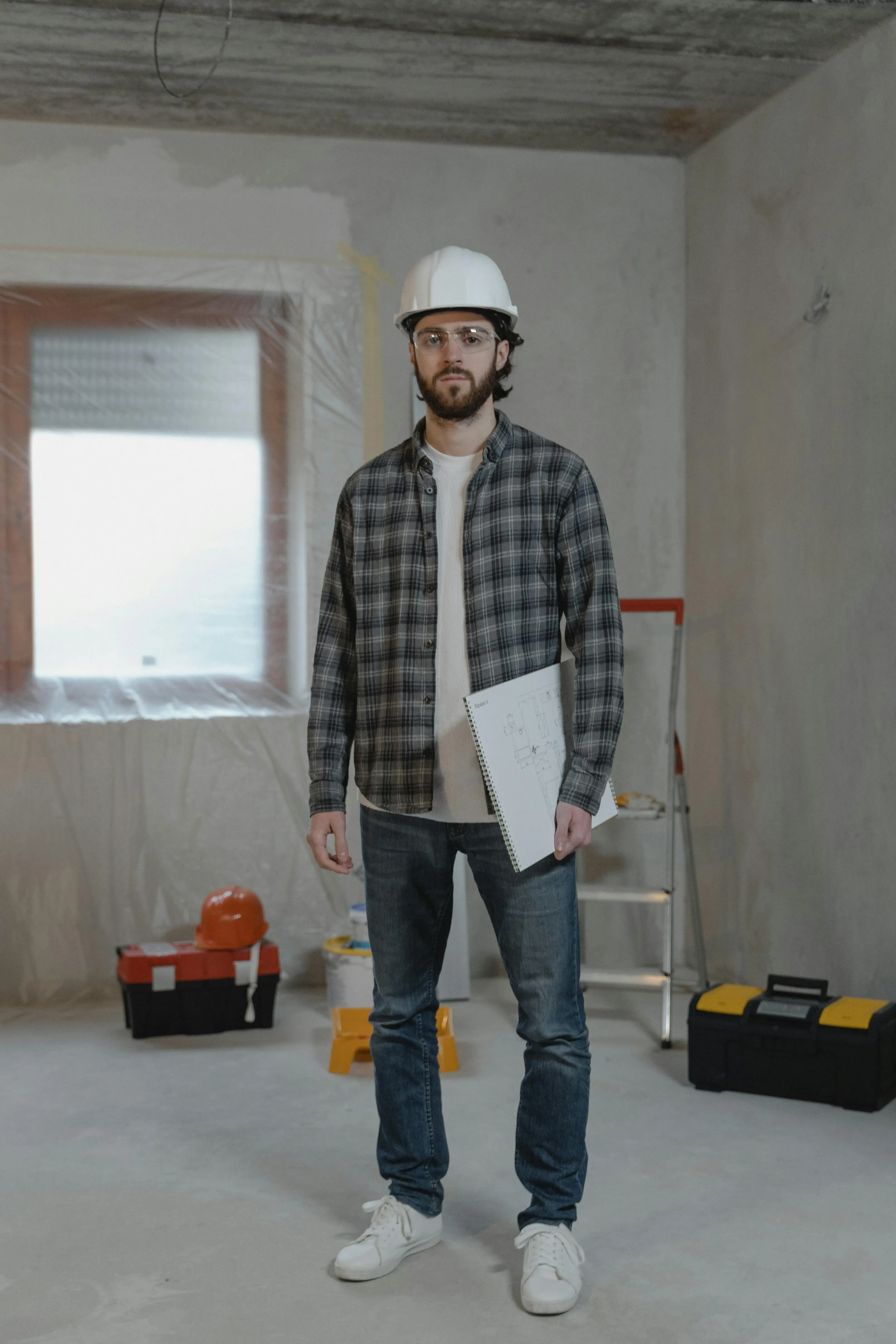 A man in a hard hat and plaid shirt stands in a room under construction, holding blueprints. Tools and a ladder are in the background. The scene conveys readiness.