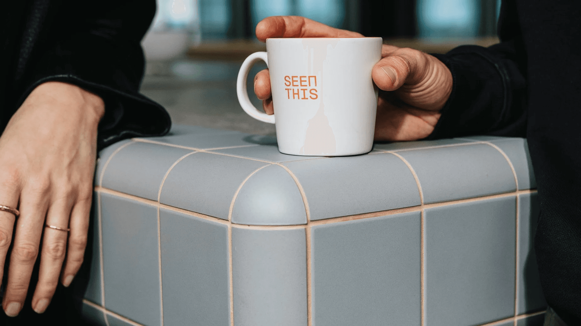 Close-up of a mug with SeenThis logo and two persons standing and talking by a bar