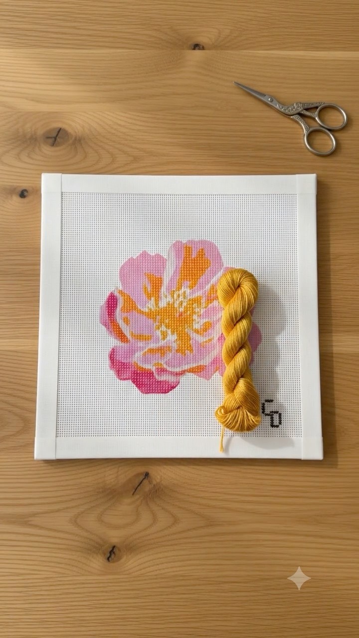 A hand-stitched needlepoint canvas of a pink and orange peony rests on a light wood table, with a small skein of golden-yellow thread and a pair of embroidery scissors placed beside it.