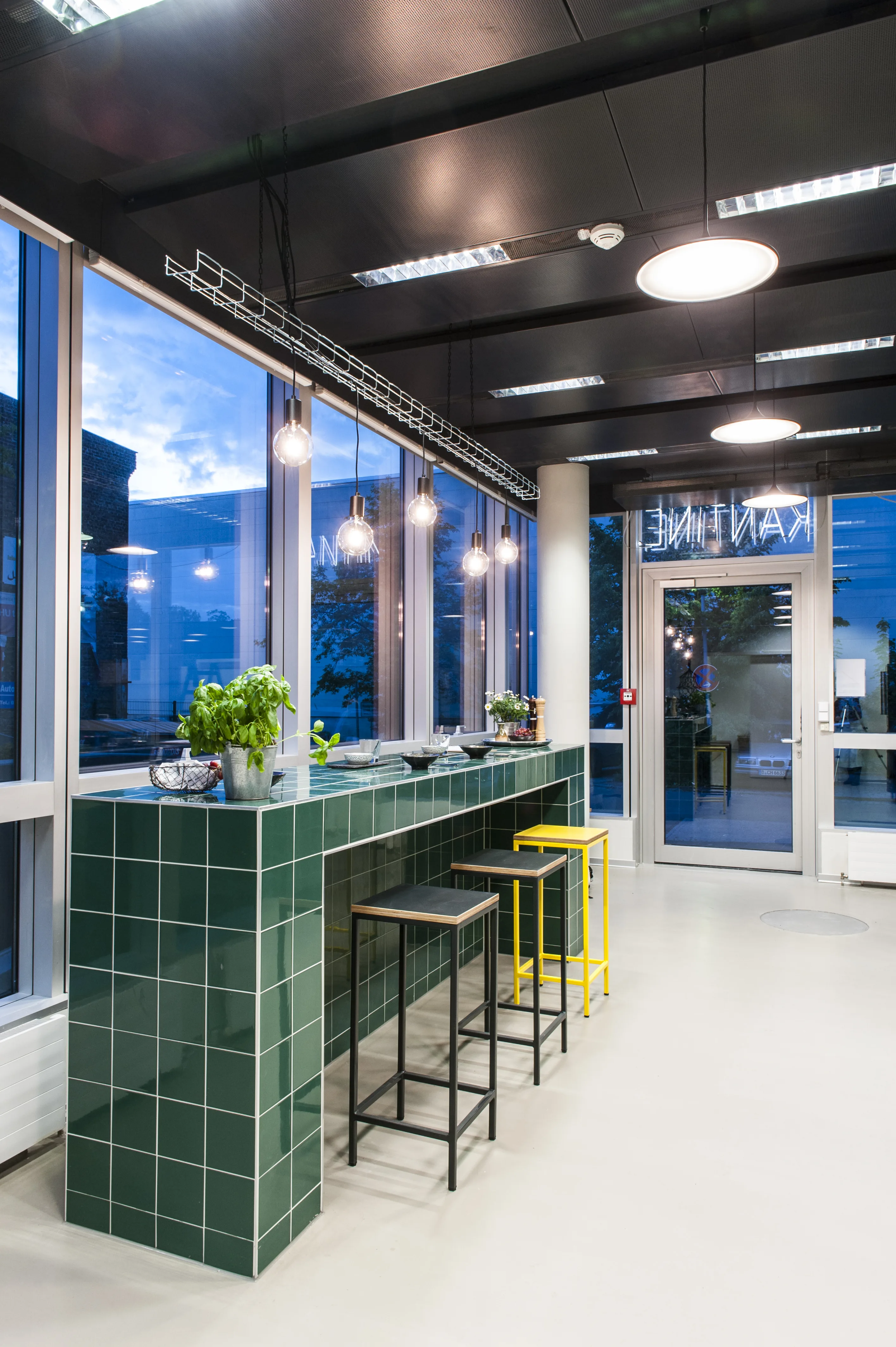Green-tiled standing counter at the entrance of KANTINE KOMTUR with yellow and black bar stools, fresh herbs and exposed filament pendant lights in front of large windows.