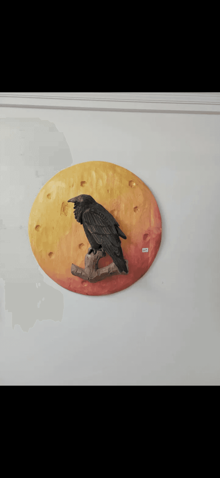 Sculpture murale d’un corbeau noir posé sur une branche devant un disque rond jaune orangé et rouge.
