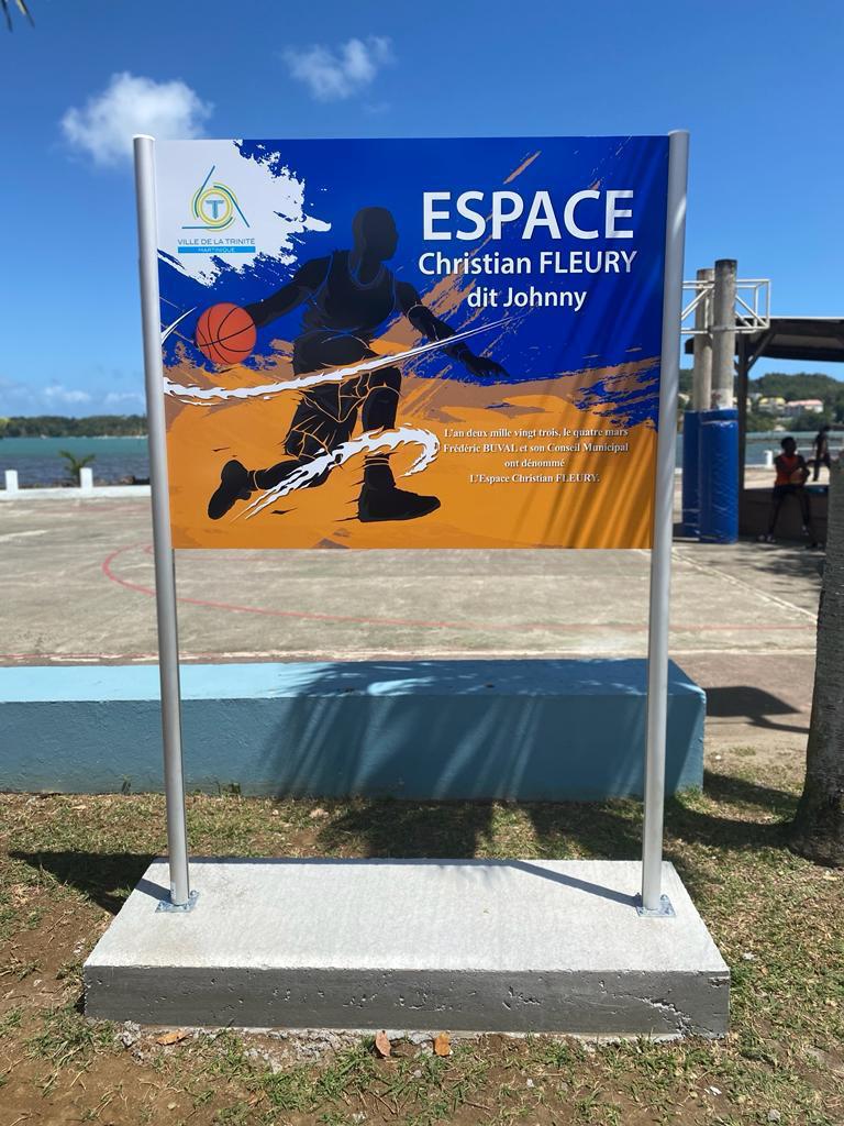 Une enseigne lumineuse représentant la silhouette d'un joueur de basket-ball sur fond bleu et orange. Le texte indique « ESPACE Christian FLEURY dit Johnny ». Journée ensoleillée.