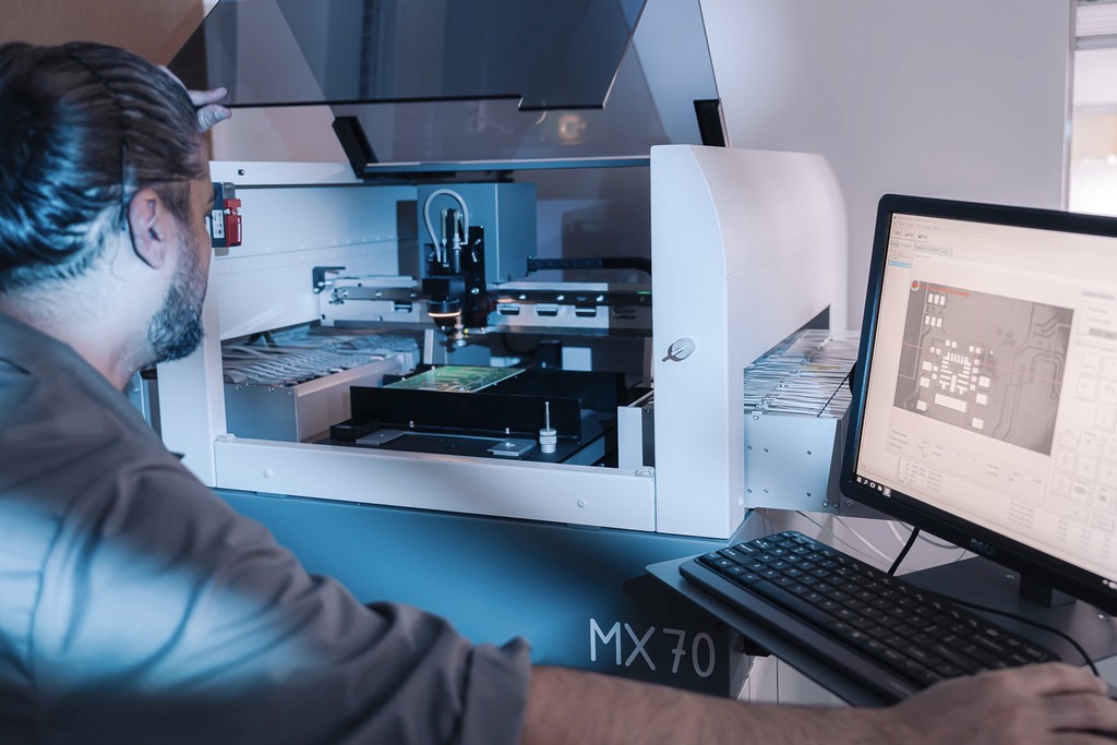 A man, viewed from behind with his head and shoulder visible, watches a machine produce a circuit board. One of his hands rests on a partially visible PC.