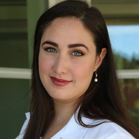A woman with long dark hair smiles at the camera, wearing a white shirt in front of a green background.
