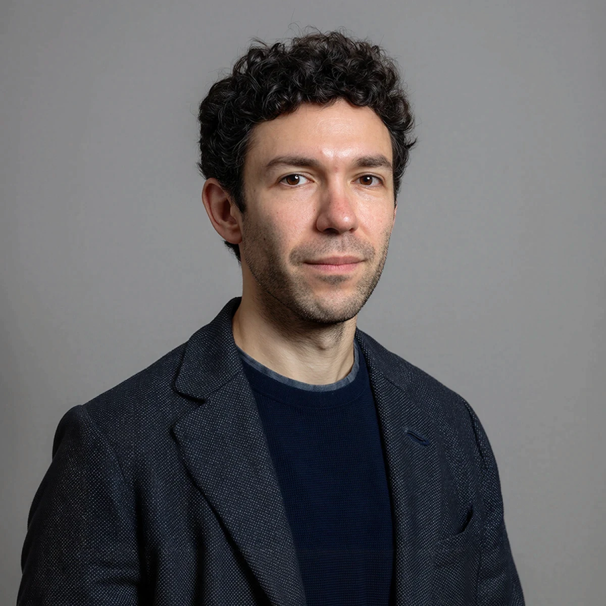 A young man with curly dark hair, wearing a black t-shirt and blazer, poses against a plain background.