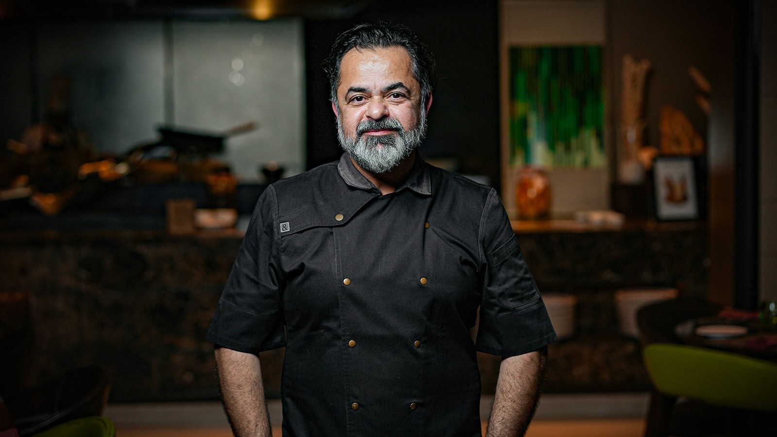 A smiling person with curly hair sits at a desk, looking at a laptop against a dark background.