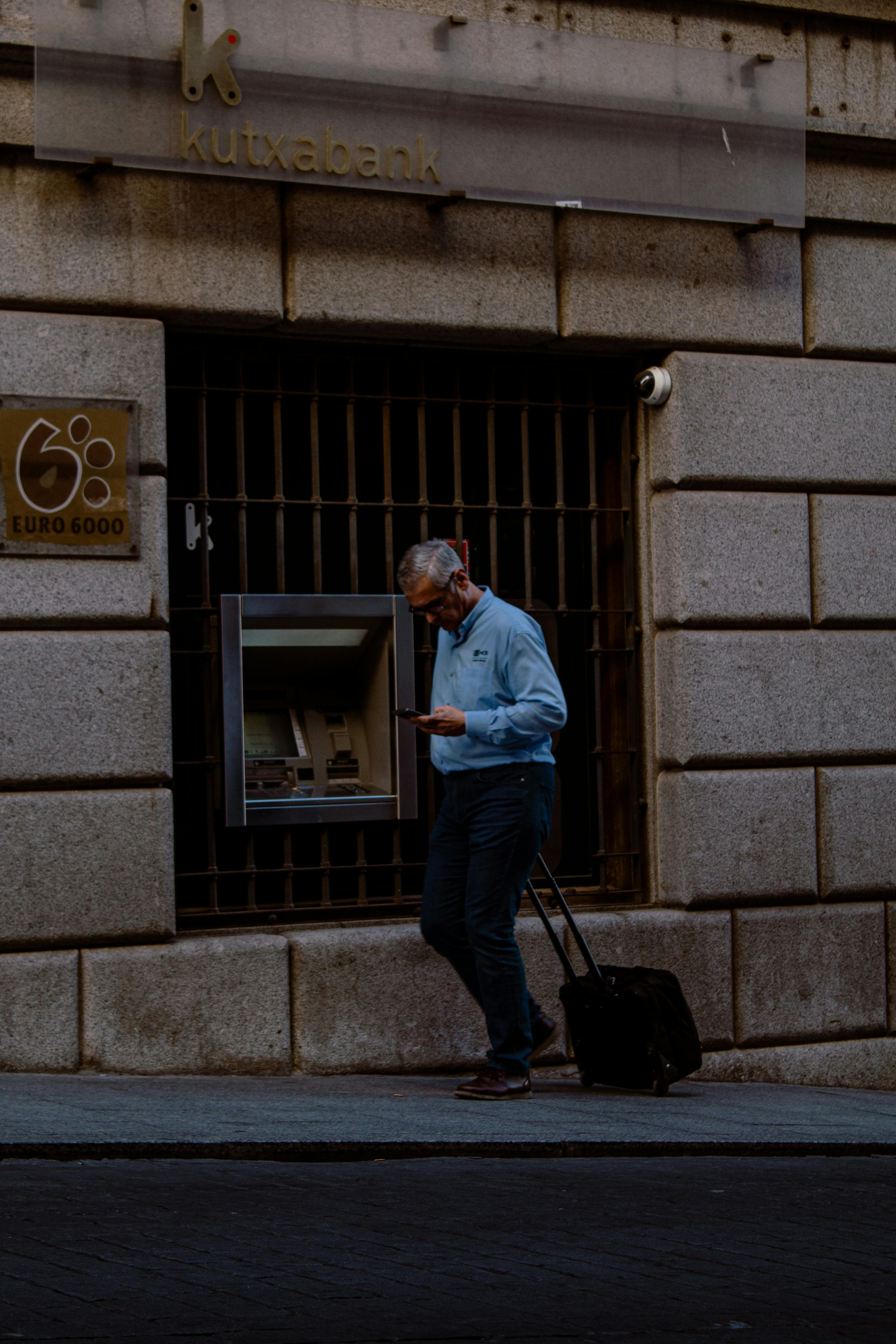 A man in a blue shirt stands at an outdoor ATM attached to a stone building, pulling a black wheeled suitcase, while checking his smartphone.