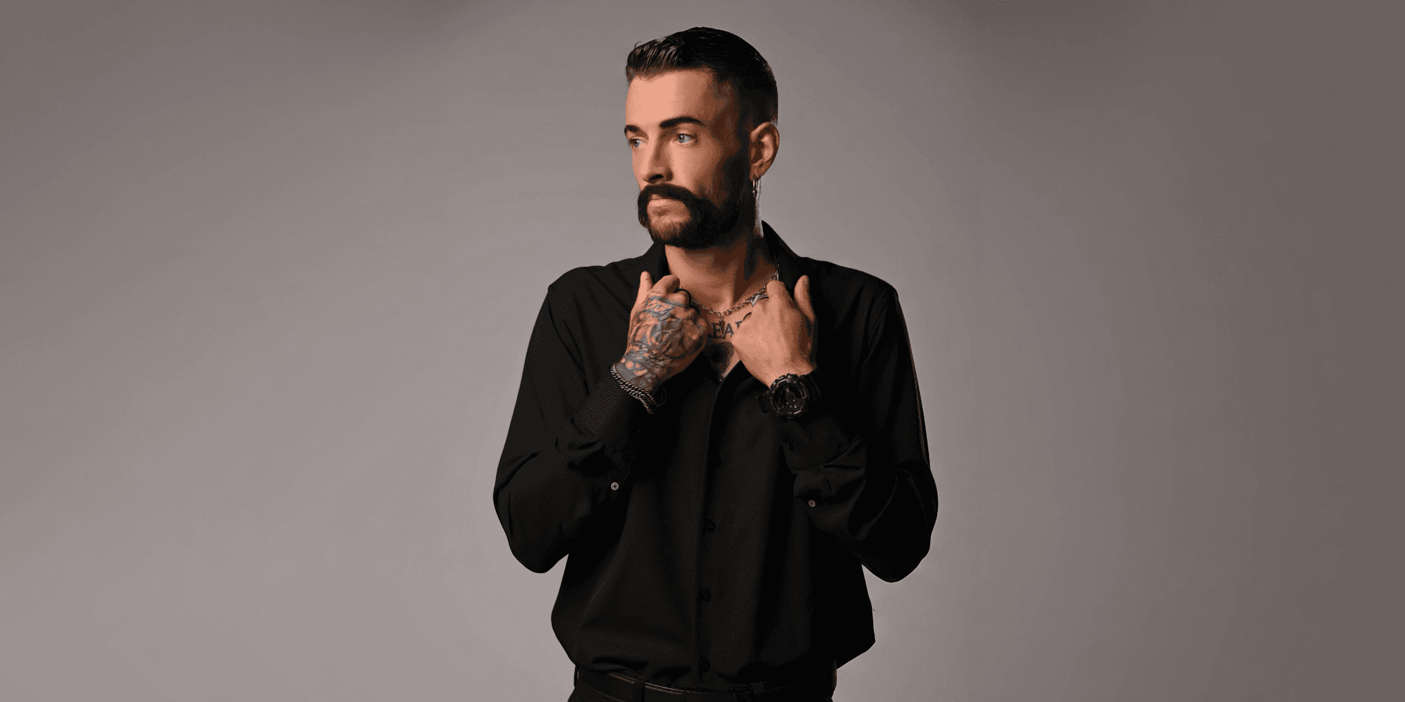 Tattooed man in black button-up shirt posing against neutral studio backdrop, looking to the side.