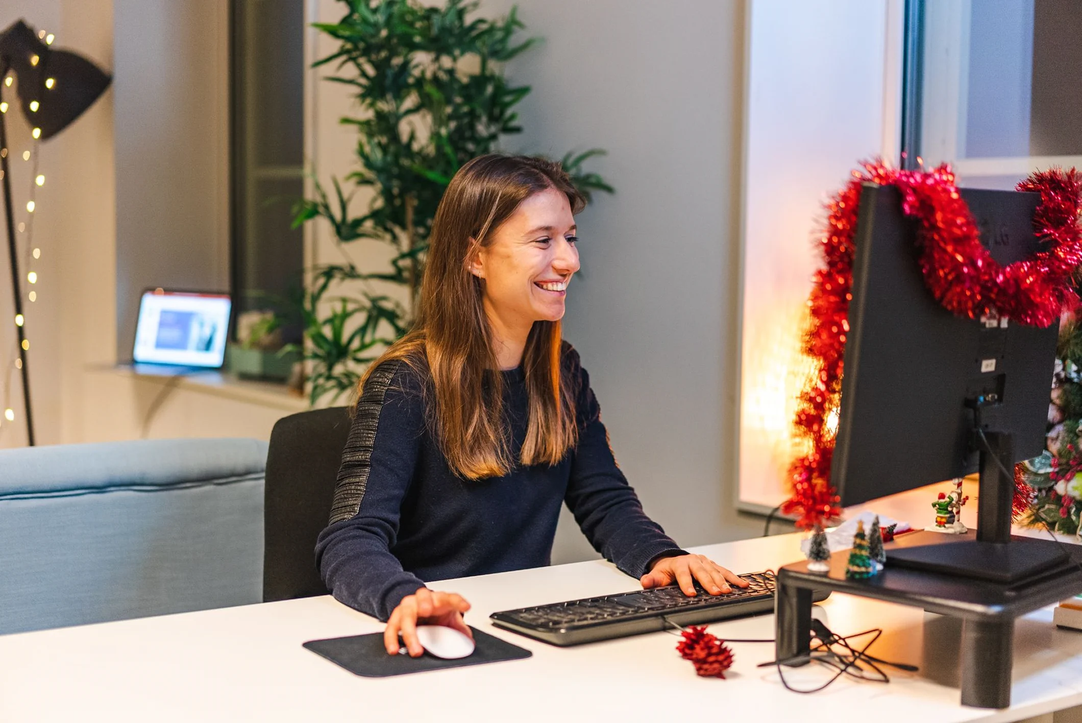 A smiling young woman with long brown hair sitting at a desk and working on a computer. She is wearing a dark blue long-sleeved sweater with textured shoulder details. Her right hand is on a white mouse and her left hand is on a black keyboard. The background features a large green indoor plant and a small, dimly lit festive red tree on the desk.