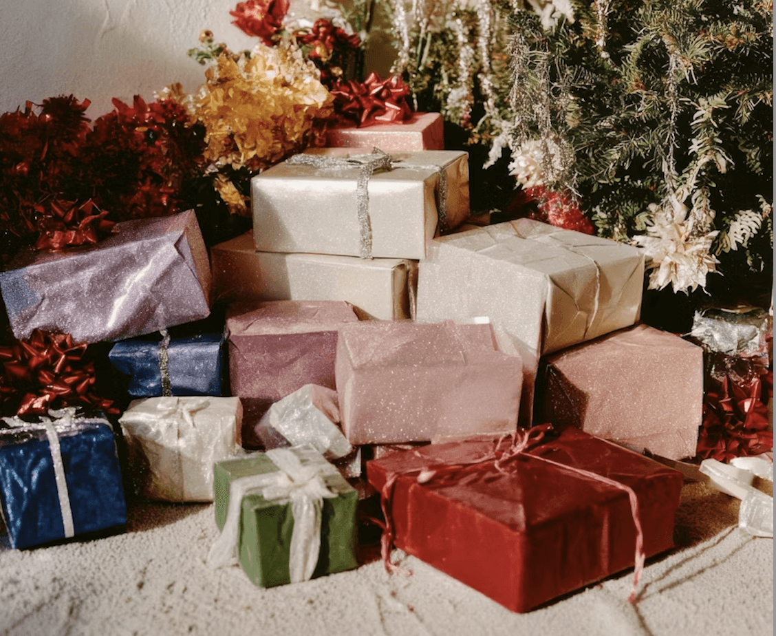 Pile of wrapped holiday gifts in assorted colors arranged beneath a decorated Christmas tree