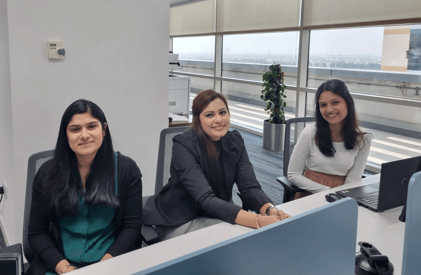 Three women sit at a modern office reception desk, smiling towards the camera with large windows in the background.