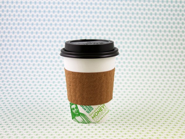 A disposable coffee cup with a black lid and a brown paper sleeve sits on a light green patterned surface. The sleeve has a green and white design featuring two faces and the words “Give back” and “Money!”. The cup is positioned slightly off-center, facing forward.
