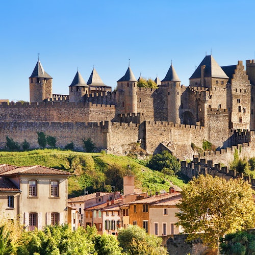 A historical castle with multiple towers overlooks a village with colorful houses and green foliage under a clear blue sky.