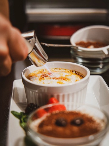 A chef torching a crème brûlée, next to a tiramisu dessert. The image shows the preparation of two desserts.