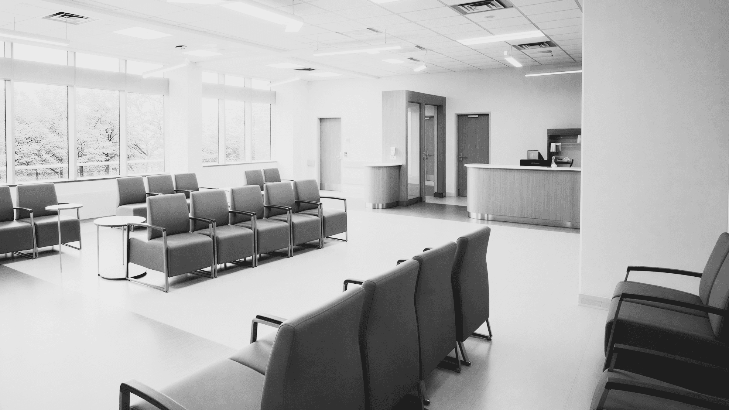 A black and white photo of a modern waiting room with teal chairs, large windows, and a reception desk in the background.
