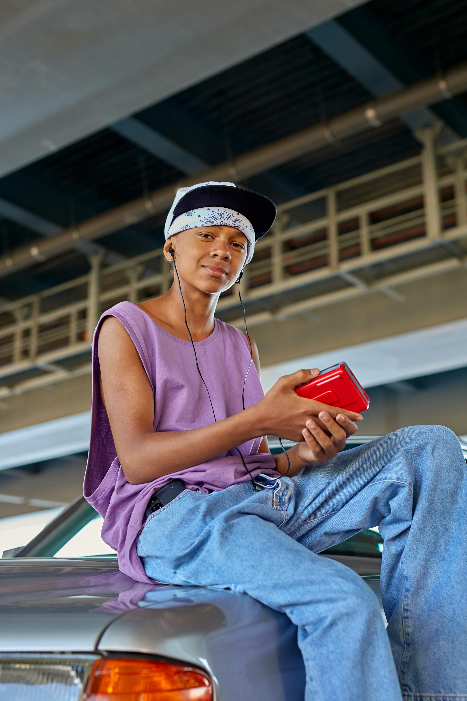 A young man wearing a purple tank top, baggy jeans, and a white bandana under a baseball cap sits on the hood of a car while holding a red portable cassette player with headphones.