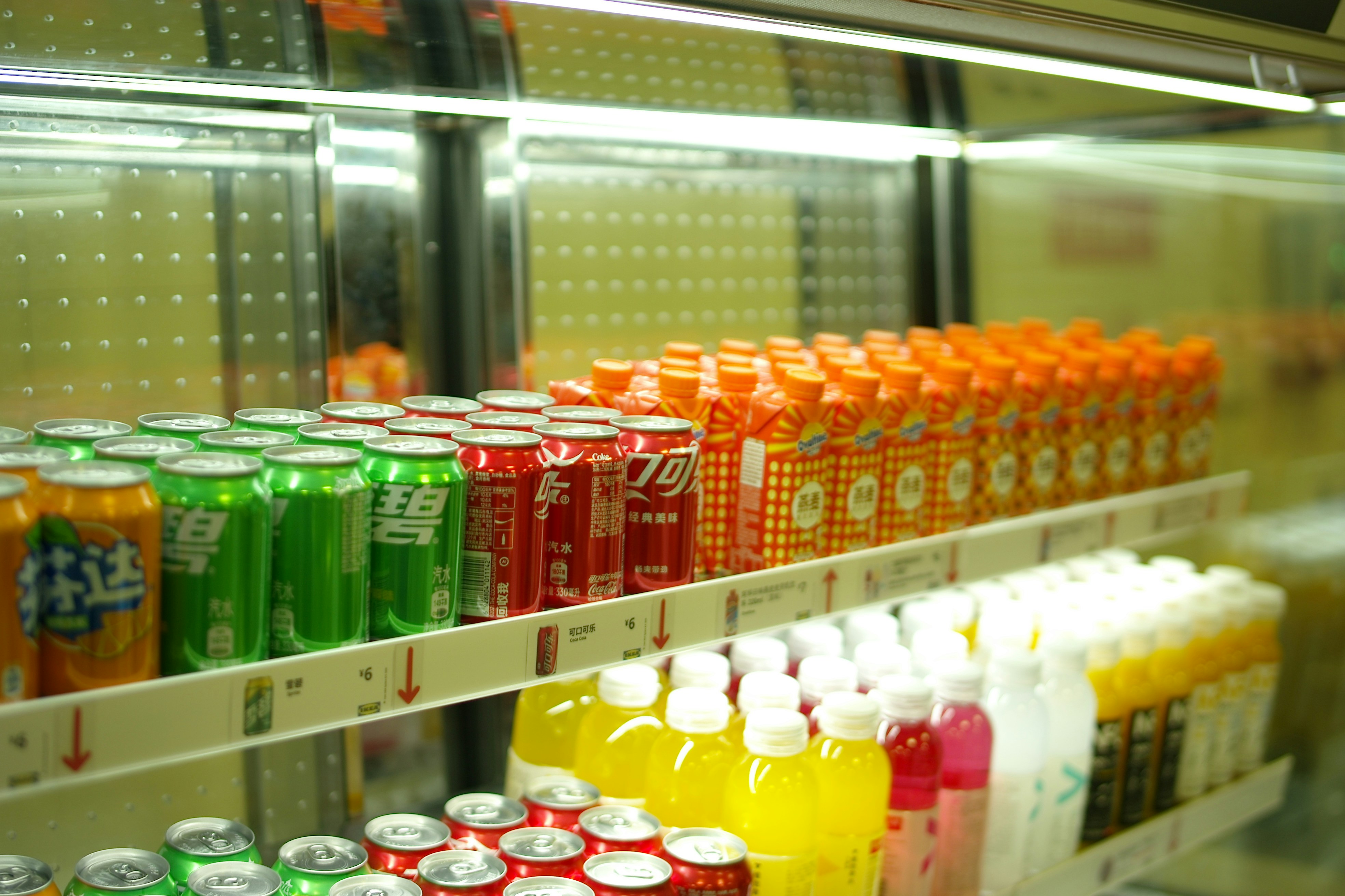 A refrigerated retail shelf filled with organized rows of colorful soft drink cans—including green and red branding—and bottled juices under bright fluorescent lighting.