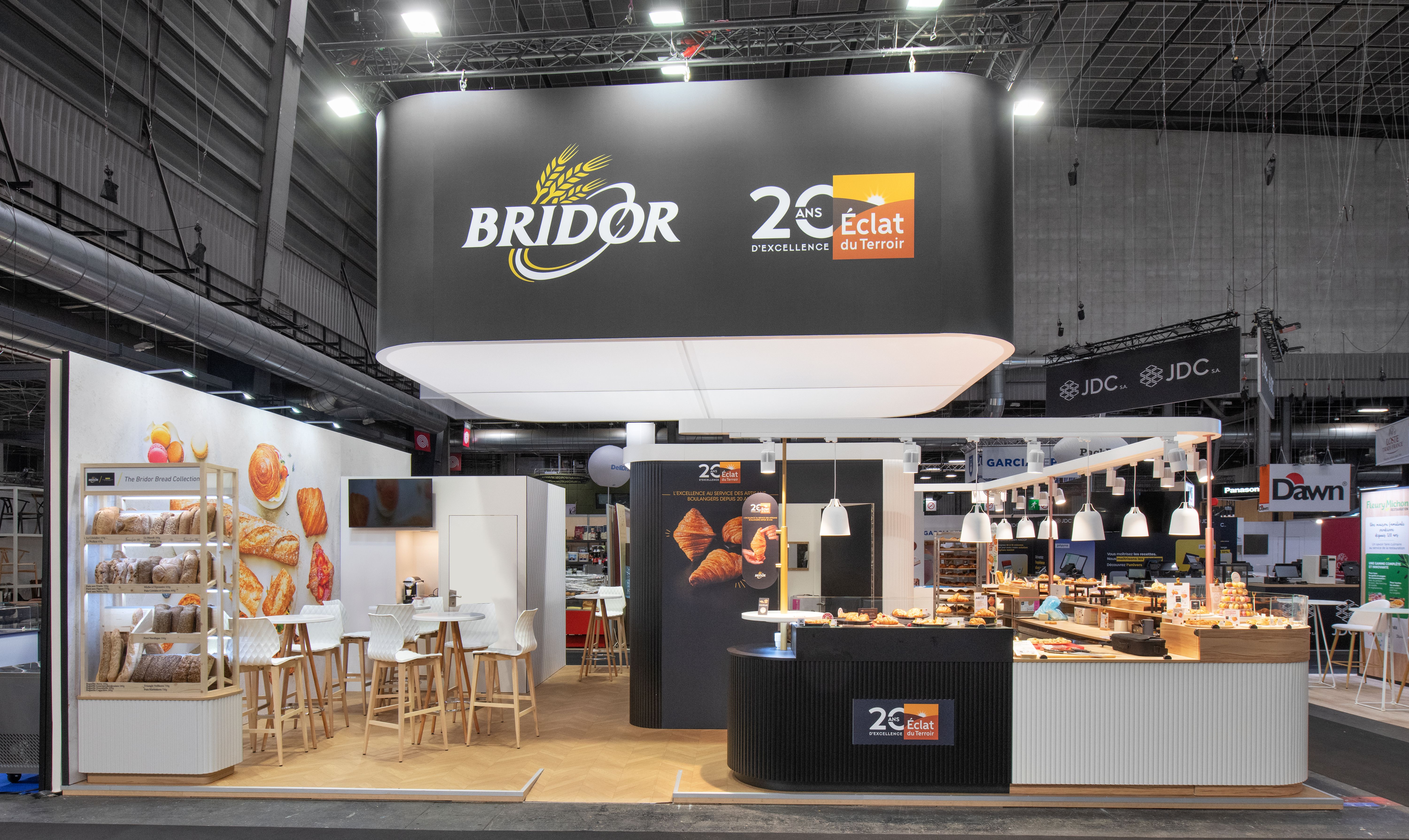 Stand Bridor Bake and Snack 2026 par Expace – vue latérale du comptoir dégustation boulangerie avec suspensions laiton, gamme produits exposée et panneaux grandes tendances boulangerie authenticité