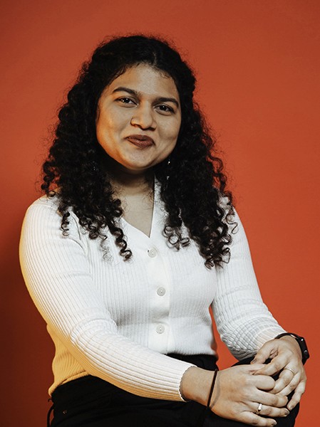 A woman with dark, curly hair sits against a solid orange-red background. She wears a white, long-sleeved button-up shirt with ribbed texture and black pants. Her hands are clasped in her lap, and she looks directly at the camera with a neutral expression.