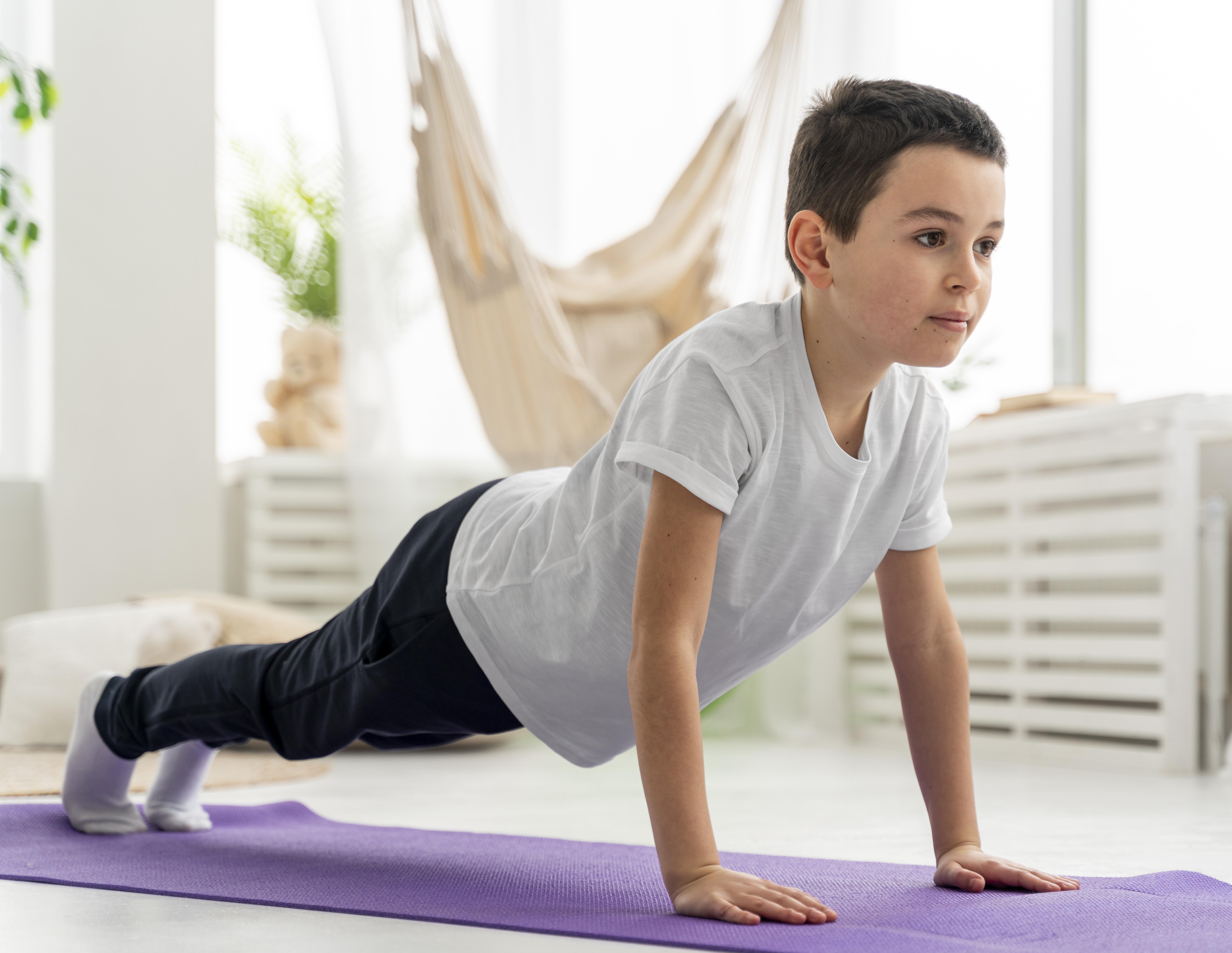 A young boy in a white T-shirt and black pants practices yoga in a plank position on a purple mat, in a bright room with a hammock and soft toys.